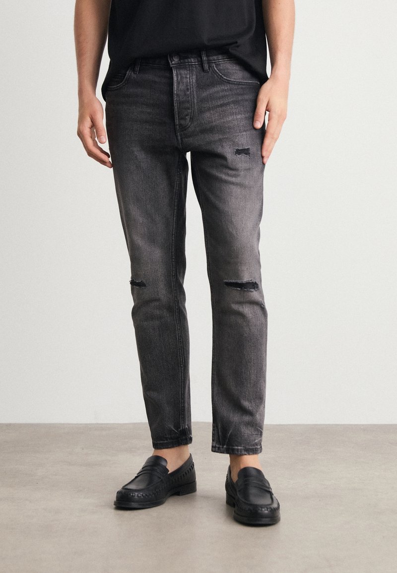Personne portant un jean noir délavé et usé ainsi que des chaussures slip-on noires, debout devant un fond clair uni.