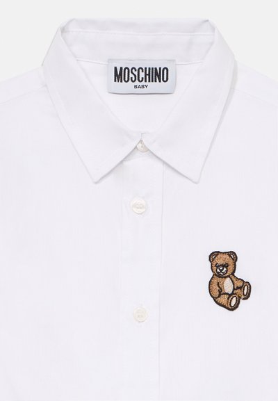 Chemise en coton blanche avec un col pointu, ornée d'un ours en peluche brodé marron sur le côté gauche et d'une fermeture à boutons à l'avant.
