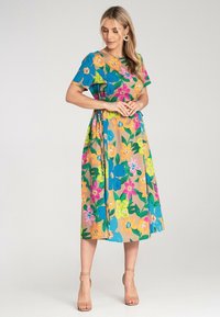 Florales Midi-Kleid mit kurzen Ärmeln in lebhaften Farben: Blau, Pink, Orange und grüne Muster auf einem beigen Hintergrund. Geknotetes Taillendetail.