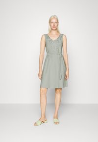 Vestido sem mangas verde-claro com bordado floral no decote e cinto de amarração. Combinado com sandálias escorregadias verde-claro. Tecido suave.