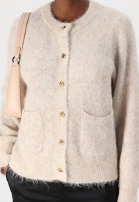 Béžový pletený cardigan s kulatým výstřihem, zlatými knoflíky a dvěma předními kapsami. Měkká, nadýchaná textura s lehce přeřazeným střihem.