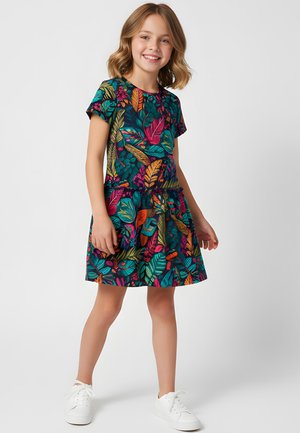 Uśmiechnięta młoda dziewczyna stojąca, ubrana w sukienkę z krótkim rękawem w kolorowe tropikalne liście oraz białe trampki na jednolitym tle.