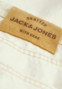 Beiger Stoff mit einem braunen, strukturierten Lederetikett. Das Etikett trägt den Text „CRAFTED JACK & JONES WITH CARE“ in Dunkelbraun.