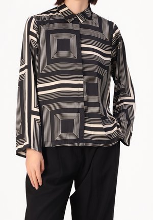 Blouse à manches longues noire et beige avec des motifs géométriques carrés portée avec un pantalon noir, main reposant le long du côté.