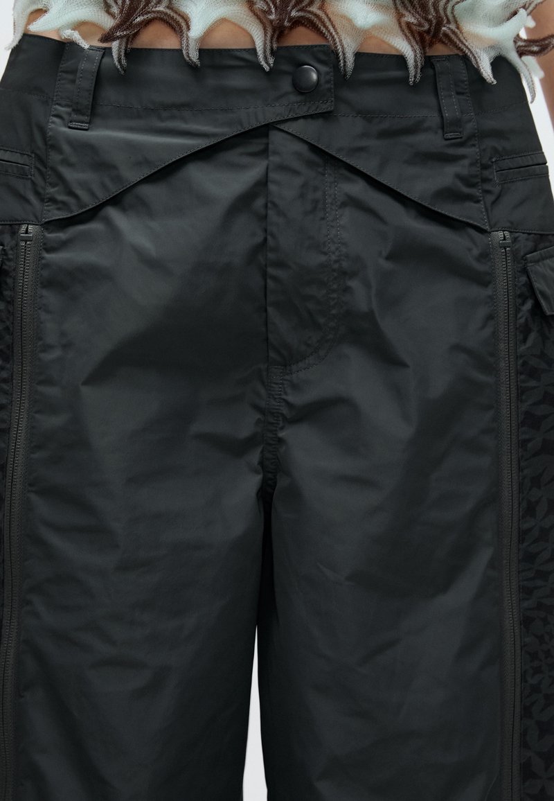 Svarta cargobyxor med dragkedja och tryckknappsstängning. Materialet är slätt med ett subtilt strukturerat mönster på sidorna av fickorna.