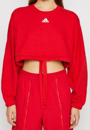 Femme portant un sweat-shirt court rouge Adidas et un pantalon rouge assorti avec taille à cordon de serrage et poches zippées.