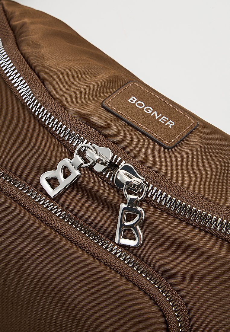 Braune Nylon-Tasche mit silbernen Beschlägen, ausgestattet mit einem auffälligen Reißverschluss, einem Logo-Patch mit "BOGNER" und zusätzlichen Reißverschlüssen. Strukturierte Oberfläche.