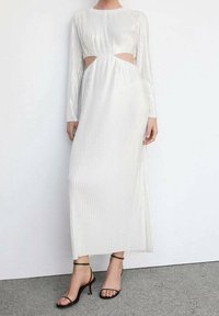 Robe blanche à manches longues avec des découpes sur les côtés, dotée de plis verticaux et d'un design fluide ; associée à des sandales à talons noirs.
