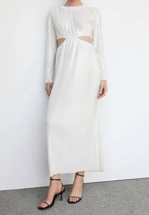 Robe blanche à manches longues avec des découpes sur les côtés, dotée de plis verticaux et d'un design fluide ; associée à des sandales à talons noirs.