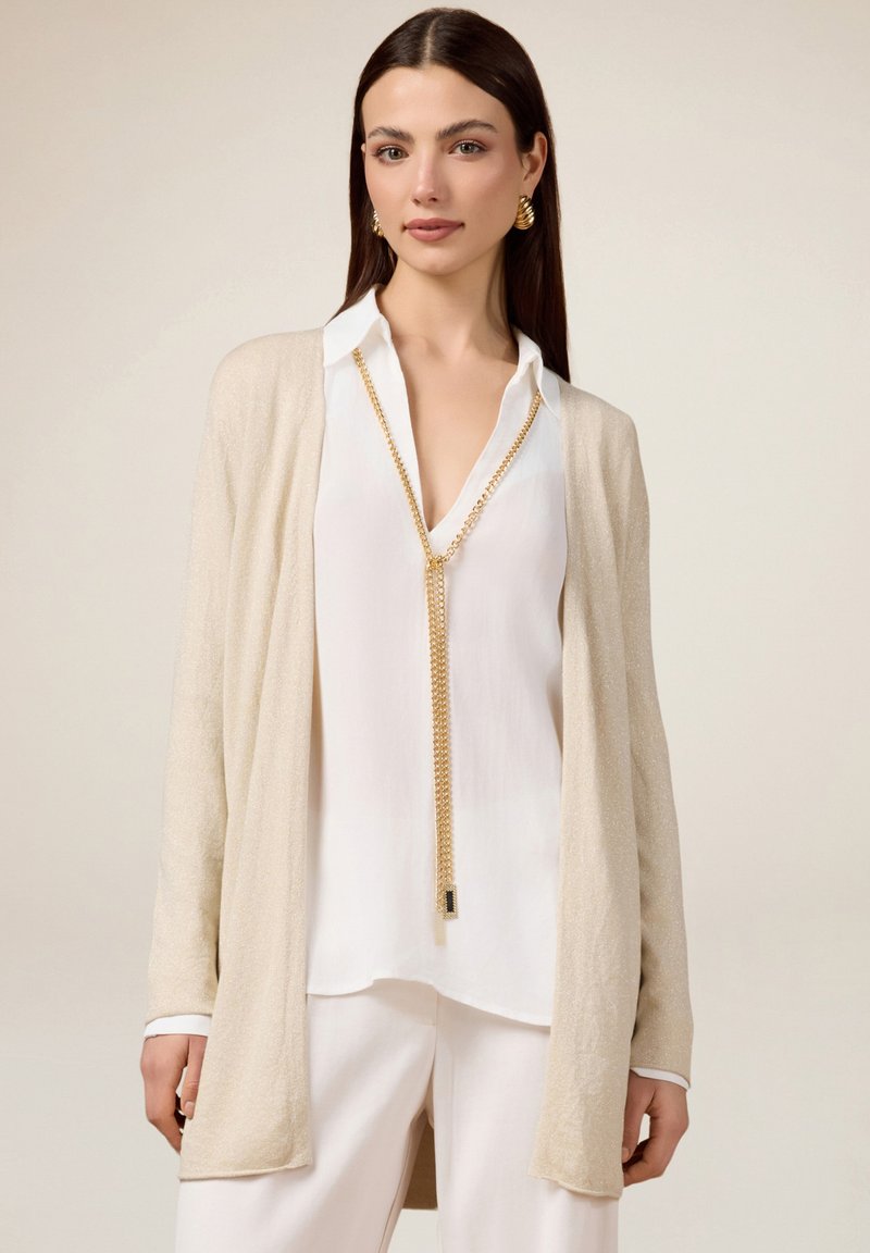 Cardigan long beige avec une texture douce sur une blouse blanche à col. Présente un collier en chaîne dorée et un pantalon blanc ample.