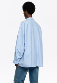Lichtblauwe oversized shirt van gladde stof, met een klassieke kraag en lange mouwen met knoopmanchetten. Eenvoudig ontwerp, geen patronen.