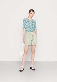 Top floral de manga curta em azul e branco, combinado com calções de cintura alta em verde claro com um detalhe de laço. Usado com sandálias brancas e uma mala.