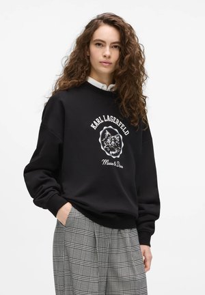 Ung kvinde med langt krøllet hår iført sort Karl Lagerfeld sweatshirt med kattegrafik og grå ternede bukser, hænderne i lommerne.