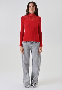 Roter, gerippter Rollkragenpullover mit transparenten horizontalen Streifen auf der Brust, kombiniert mit hellgrauen, weit geschnittenen Jeans und metallic-farbenen spitzen Schuhen.