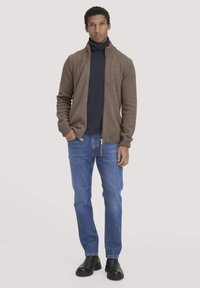 Brauner Strickcardigan mit hohem Kragen, Reißverschluss und gerippten Bündchen, getragen über einem dunklen Rollkragenpullover, kombiniert mit blauen Jeans und schwarzen Stiefeln.