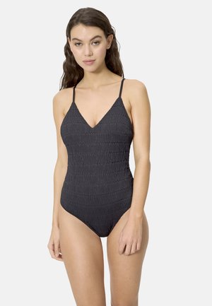 Mujer con un traje de baño de una pieza negro texturizado y tirantes finos, de pie contra un fondo claro y simple.