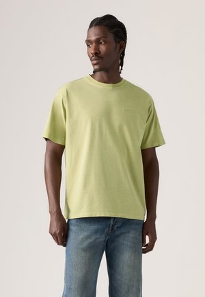 Homme aux cheveux tressés portant un t-shirt vert clair à manches courtes et un jean bleu, regardant légèrement sur le côté sur un fond uni.