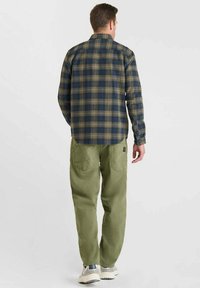 Man in een blauw en groen geruit overhemd met lange mouwen, olijfgroene broek en lichtgekleurde sneakers, staande met de rug naar de camera gekeerd.