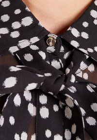 Blouse noire à pois blancs, avec un col noué et un bouton argenté. Fabriquée en tissu léger avec une texture douce.