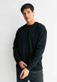 Černá mikina s kulatým výstřihem v uvolněném střihu a dlouhými rukávy, vyrobená z měkké látky. Má žebrované manžety a lemování, v kombinaci s tmavými džíny.