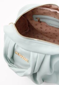 Vaaleansininen nahkalaukku, jossa on poimuinen muotoilu. Sisäosassa on vaaleanbeige vuori, jossa on toistuva brändilogotunnus, sivupussukka ja kultaiset yksityiskohdat.