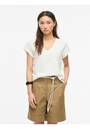 Kvinde iført hvid V-hals kortærmet top, tan-farvede shorts til knæet med hvid snor og sort armbånd, stående med den ene hånd i lommen.