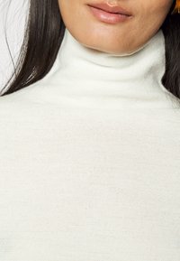 Kobieta nosząca biały sweter z dzianiny z wysokim kołnierzem, z długimi ciemnymi włosami, widoczna na jednolitym tle.