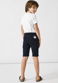 Marina bomullsshorts med mjuk midja, två bakfickor och en etikett. Bärs med en vit t-shirt och sandaler, sedd bakifrån.