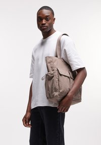 Sac fourre-tout beige rembourré avec une poche latérale zippée et deux poignées. Tenue par une personne portant un t-shirt blanc et un jean foncé.