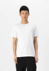 T-shirt en coton blanc avec un col rond, des manches courtes et une coupe décontractée, porté avec un pantalon cargo noir. Design minimaliste, sans motifs visibles.
