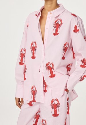 Personne portant une chemise rose pâle ample et un pantalon à l'imprimé répétitif de homard rouge, chemise partiellement déboutonnée et main dans la poche.