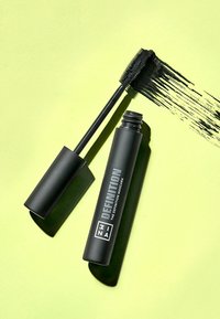 Tubetto di mascara nero con finitura opaca e testo metallico, dotato di un pennello con setole fitte, su uno sfondo verde chiaro.