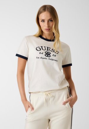 Woman wearing a white Guess t-shirt with black trim and text, paired with light-colored drawstring pants with side stripes, standing with hands in pockets.