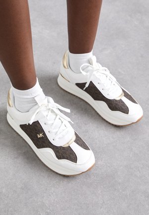 Persona che indossa sneakers bianche con pannelli marroni a motivi, dettagli dorati, calze bianche, in piedi su pavimento grigio.