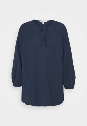 Blusa azul marino con mangas largas abullonadas, escote redondo y detalle de lazo. Presenta un tejido texturizado con pequeños puntos elevados.