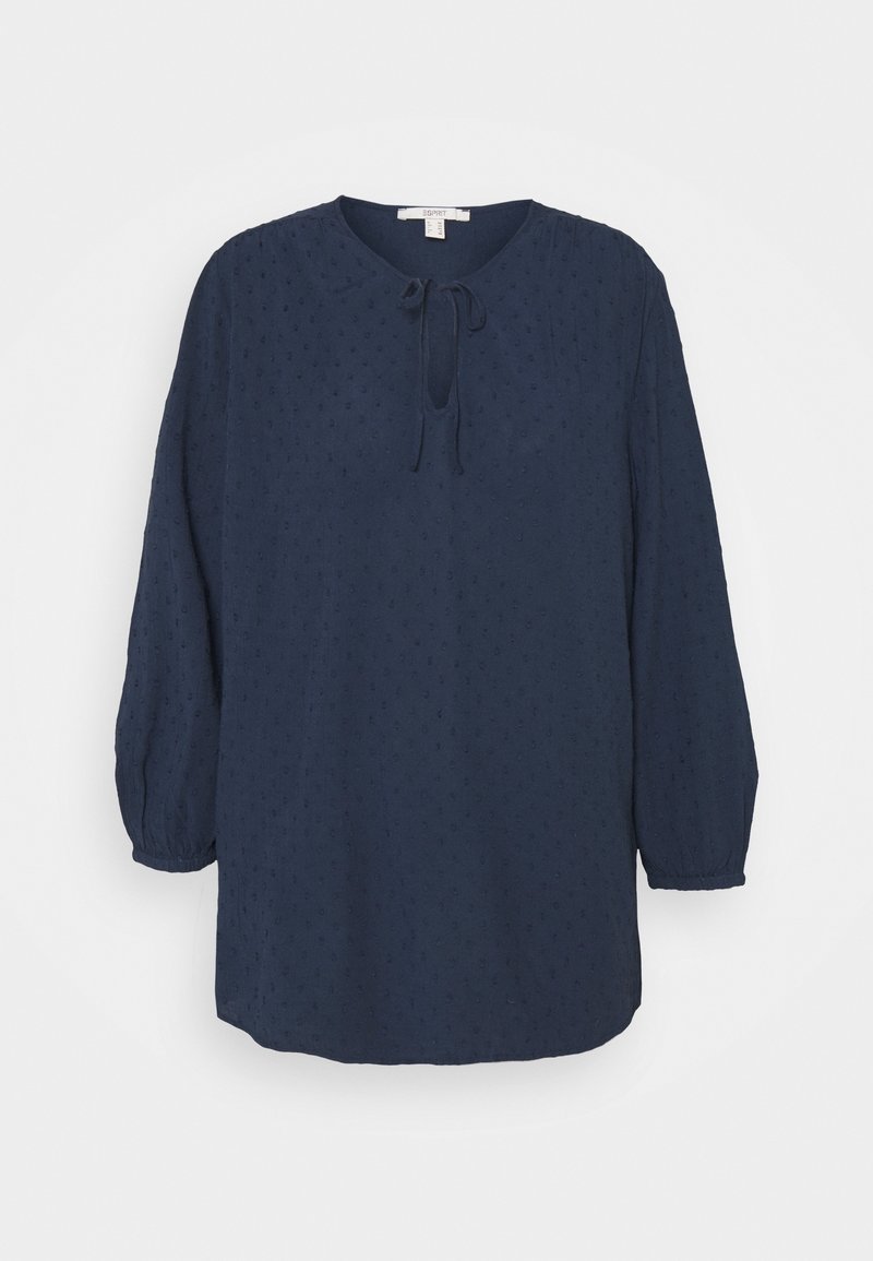 Blouse bleu marine avec manches longues bouffantes, encolure ronde et détail noué. Tissu texturé avec petits points en relief.
