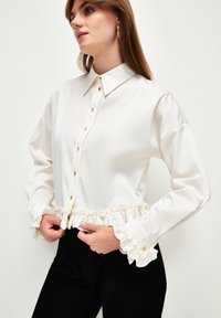Camicia bianca con colletto e polsini arricciati, dotata di bottoni dorati e tessuto liscio. Indossata con pantaloni neri.