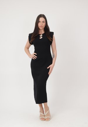 Kvinde stående med den ene hånd på hoften iført en sort midi-kjole uden ærmer og beige strop-sandaler mod en hvid baggrund.