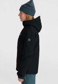 Schwarze wasserdichte Jacke mit Kapuze, strukturiertem Stoff und Reißverschlussfächern. Getragen mit einer blauen Mütze, die ein Logo auf der linken Schulter hat.