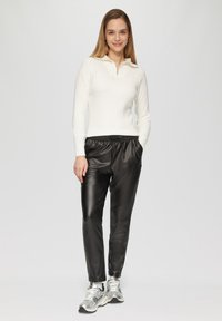 Witte geribbelde pullover met een ritssluiting bij de kraag, gecombineerd met zwarte neplederen broeken. Het model draagt zilveren sportschoenen en staat met de handen in de zakken.