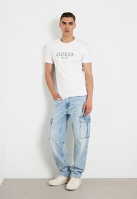 T-shirt en coton blanc avec logo vert "GUESS EST. 1981", associé à un pantalon cargo en denim bleu clair avec des poches latérales et une coupe décontractée.