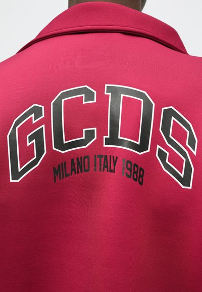 Zadnja stran rdečega jopiča z velikimi črnimi in belimi črkami "GCDS" ter manjšim besedilom "Milano Italija 1988" pod ovratnikom.