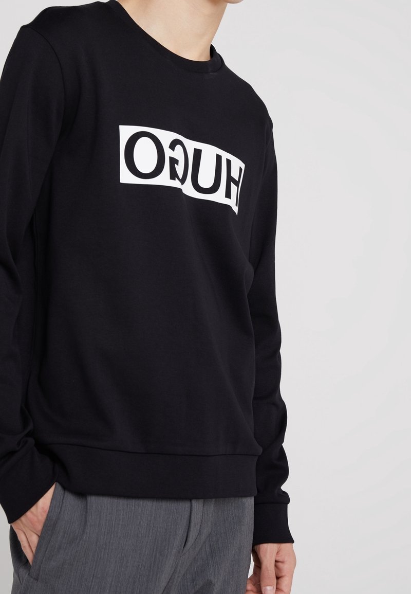 Person in einem schwarzen Sweatshirt mit einem weißen rechteckigen Patch und umgekehrtem "HUGO"-Text, kombiniert mit grauen Hosen vor einem schlichten Hintergrund.