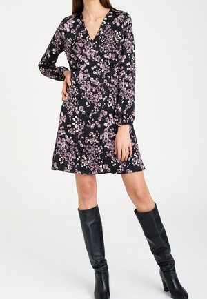Femme portant une robe à fleurs noire à col en V avec manches longues et des bottes en cuir noires montantes jusqu'aux genoux, debout avec une main sur la hanche.