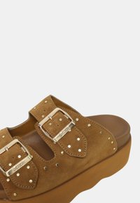 Sandal con zeppa in suede marrone, dotato di due fibbie metalliche, accenti di borchie e una suola spessa. Texture morbida e design aperto per una buona traspirabilità.