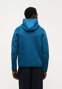 Hoodijs spilgtā zilā krāsā ar gludu tekstūru, šuvēm un melniem akcentiem pie apakšmalas un aprocēm.