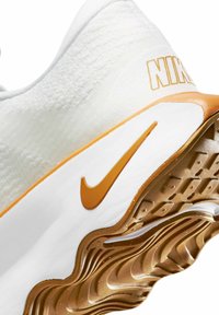 Nike tenisice s bijelim tekstilnim gornjištem i zlatnim akcentom. Potplat ima valoviti dizajn u zlatnoj boji s teksturiranim uzorcima za prianjanje.