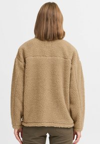 Veste en polaire beige duveteuse avec une coupe décontractée, épaules tombantes et détails de couture dans le dos. Texture douce avec un ourlet arrondi.