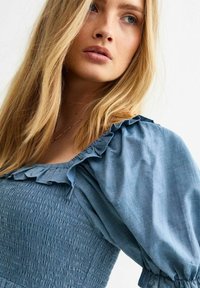Vestido azul claro con un cuerpo fruncido, escote con volantes y mangas abullonadas. La tela parece suave con una sutil textura.