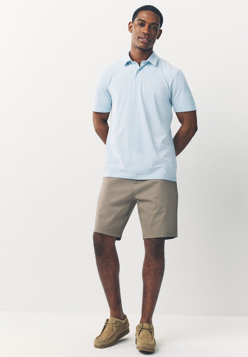 Šviesiai mėlyna polo marškeliai, trumpomis rankovėmis. Neutralūs chaki šortai. Rudos zomšos laisvalaikio avalynės su raišteliais. Paprastas dizainas, be matomų raštų ar logotipų.